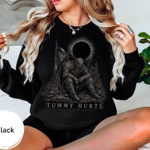 Könnte beinhalten: Ein schwarzes Langarm-Sweatshirt mit einem Grafikdruck eines in Rüstung gekleideten Ritters, der unter einer dunklen Sonnenfinsternis auf dem Boden sitzt. Der Text "TUMMY HURTS" ist in weißen Großbuchstaben unter dem Ritter aufgedruckt.