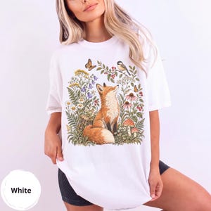Puede incluir: Una camiseta blanca con un estampado gráfico colorido de un zorro sentado entre flores silvestres, helechos, setas, un pájaro pequeño y mariposas. La ilustración incluye varios elementos botánicos en tonos naranja, verde y rojo.