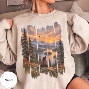 May include: A sand-colored crewneck sweatshirt featuring a graphic print of a bear sitting on a rocky outcrop overlooking a lake, pine trees, and snow-capped mountains at sunset, with birds flying in the orange and yellow sky.