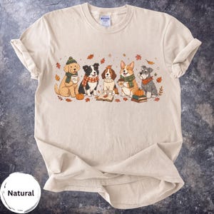 Könnte beinhalten: Ein sandfarbenes T-Shirt mit einer grafischen Illustration von fünf Hunden, die Schals und Mützen tragen, umgeben von Herbstblättern, Kürbissen und Büchern. Die Hunde sind in verschiedenen Posen dargestellt, einige halten Tassen mit der Aufschrift PSL.