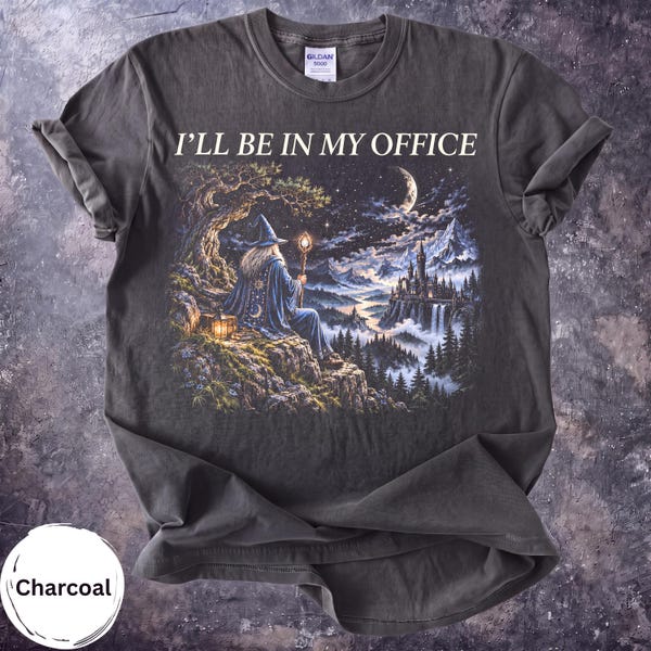 May include: A charcoal gray t-shirt featuring the text I'LL BE IN MY OFFICE above a graphic of a wizard in a blue robe and hat holding a glowing staff. The scene depicts a wizard sitting on a cliffside overlooking a castle and mountains.