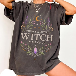 Könnte beinhalten: Ein anthrazitfarbenes Kurzarm-T-Shirt mit einem Grafikdruck und dem Text "THERE'S A LITTLE WITCH IN ALL OF US". Das Design zeigt eine Mondsichel, florale Zweige und einen violetten Kristall, die in einem symmetrischen botanischen Rahmen angeordnet sind.