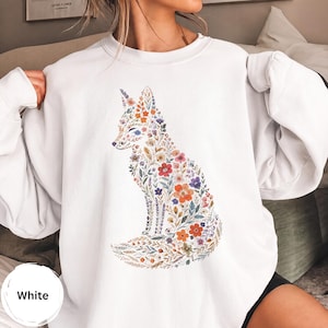 Peut inclure: Un sweat-shirt blanc à col rond arborant un motif central représentant une silhouette de renard composée entièrement de fleurs sauvages colorées. Le design floral inclut des tons orange, rouge, violet, bleu et vert, formant un renard botanique.