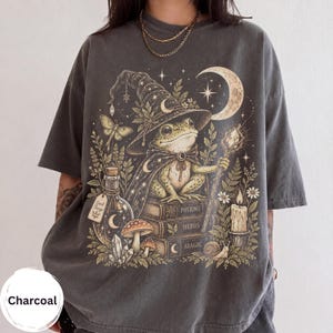 Könnte beinhalten: Ein anthrazitfarbenes T-Shirt mit einem Grafikdruck eines Frosches, der einen Zaubererhut und einen Umhang trägt und auf einem Stapel Bücher mit der Aufschrift Potions, Herbs und Magic sitzt. Das Design zeigt einen Halbmond, eine Kerze, Pilze und eine Flasche.