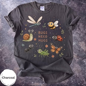 May include: A charcoal grey crew neck t-shirt featuring a graphic print of various cartoon insects including a dragonfly, bee, snail, ant, grasshopper, ladybug, and butterfly, with the text "BUGS NEED HUGS" printed in the centre in a light colour.