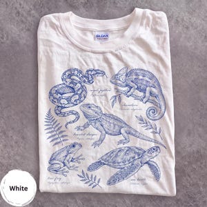Könnte beinhalten: Ein weißes T-Shirt mit einer blauen Tuschezeichnung eines Königspythons, Chamäleons, einer Bartagame, eines Laubfroschs und einer Meeresschildkröte. Jedes Tier ist mit seinem deutschen und wissenschaftlichen Namen in Schreibschrift sowie Farnblättern versehen.
