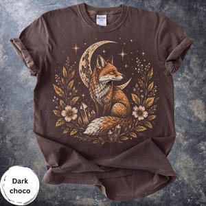 Peut inclure: Un t-shirt marron chocolat foncé avec une impression graphique représentant un renard assis devant un croissant de lune, entouré d'éléments botaniques, d'étoiles et de cristaux. L'étiquette de la marque Gildan est visible au col.