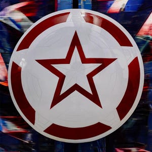 May include: A circular shield featuring a prominent central five-pointed star in white with a red outline. The design includes a wide red outer ring and a white inner ring, with geometric paneling details embossed across the surface of the shield.