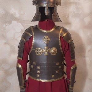 Op de afbeelding: Een harnas van donker metaal met een helm voorzien van twee verticale vleugelachtige kammen, een borstplaat versierd met drie cirkelvormige goudkleurige kruisemblemen, gesegmenteerde schouderstukken en onderarmplaten, gedragen over een lange rode tuniek.