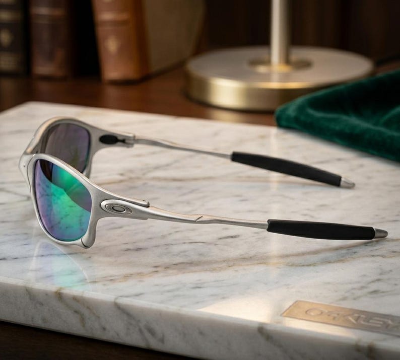 May include: Silver-framed sunglasses with iridescent lenses. The sunglasses have a sleek, modern design with black temple tips. The lenses reflect a spectrum of colors, including green and blue. The Oakley logo is visible on the frame.