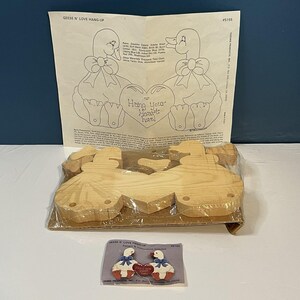 May include: A craft kit featuring unfinished wooden cutouts of two geese holding a heart, accompanied by a printed instruction sheet titled Geese N' Love Hang-Up #5155. The kit includes a small color reference card showing the finished painted design.