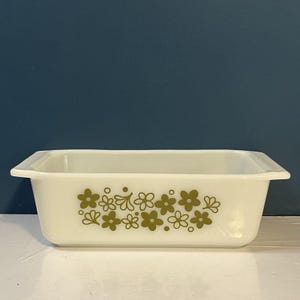 Op de afbeelding: Een rechthoekige witte glazen ovenschaal met geïntegreerde handgrepen aan de zijkanten. De buitenkant is voorzien van een herhalend patroon van olijfgroene gestileerde bloemen en kleine cirkels in het midden van de voorzijde.
