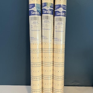 May include: Three rolls of Sanitas wallpaper, each wrapped in clear plastic. The wallpaper features a cream background with a repeating pattern of thin horizontal lines, small dots, and delicate floral borders in green and pink hues.