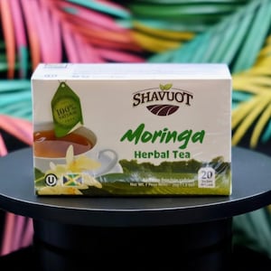 May include: A white box of Shavuot Moringa Herbal Tea with a green label that says "100% Natural". The box has a picture of a cup of tea and the Jamaican flag. The box contains 20 tea bags.