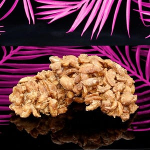 May include: Close-up of two clusters of caramel-covered nuts, likely a snack or treat. The nuts are a light brown color, with a glossy coating. The background is black with bright pink palm fronds.