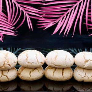 May include: A stack of four light-colored, round cookies with cracked surfaces. The cookies are arranged on a black surface, with pink palm fronds in the background. The cookies appear to be freshly baked.