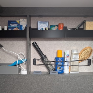 May include: A dark grey bathroom medicine cabinet with open doors, featuring black storage shelves holding boxes of Universal Plasters and Nasa Rhathiol. The lower section contains a hairbrush, Nivea sunscreen, toothpaste, and a hair styling brush.