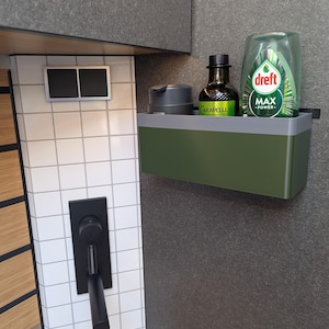 May include: A green and grey wall-mounted shelf holding a bottle of olive oil, a grey container, and a bottle of Dreft Max Power detergent. The shelf is mounted on a dark grey wall next to white tiled wall with a black tap.