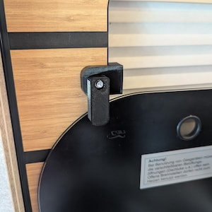 Pode incluir: Um mecanismo de fecho em plástico preto impresso em 3D montado num painel com textura de madeira e riscas pretas horizontais. O fecho possui um parafuso metálico prateado central e fixa um painel preto com uma etiqueta de aviso branca.