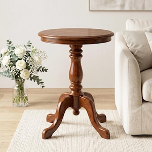 Puede incluir: Una mesa auxiliar redonda de madera marrón oscuro con base de pedestal y tres patas curvas. La mesa tiene una superficie lisa y pulida y mide aproximadamente 61 cm de altura. Está colocada sobre una alfombra color crema.