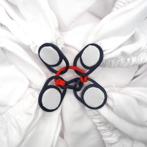 Puede incluir: Cierre decorativo de cuatro partes con un anillo central rojo y anillos exteriores azul marino. Cada anillo azul marino sostiene una almohadilla circular blanca, sobre un fondo de tela blanca.