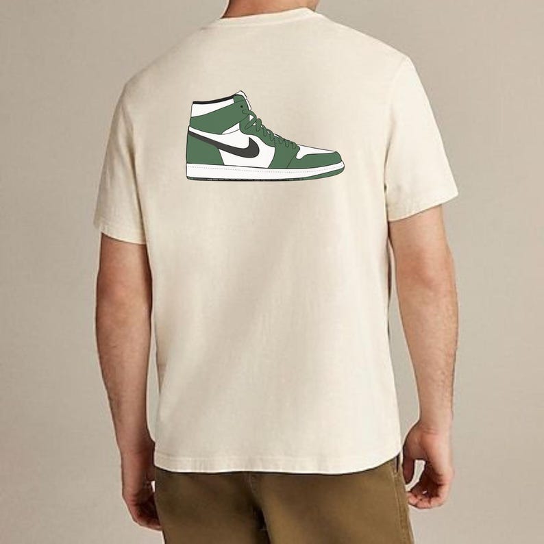 May include: A cream-colored short-sleeve t-shirt featuring a graphic illustration on the back. The graphic depicts a high-top sneaker in white, dark green, and black colors, shown from a side profile view with laces and a black swoosh logo.