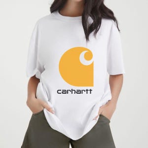 May include: A white short-sleeved t-shirt featuring a large yellow circular logo with a crescent cutout on the chest. Below the graphic, the word carhartt is printed in a bold, black, sans-serif font. The garment has a crew neckline.