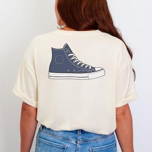 May include: A cream-colored short-sleeve t-shirt featuring a graphic print of a blue high-top sneaker on the back. The sneaker illustration includes white laces, a white rubber toe cap, a white sole, and a circular patch detail on the side.