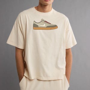 May include: A cream-colored short-sleeve t-shirt featuring a centered graphic illustration of a low-top sneaker. The sneaker design displays a light beige upper, a white swoosh logo, dark red laces, and a solid gum-colored rubber sole.