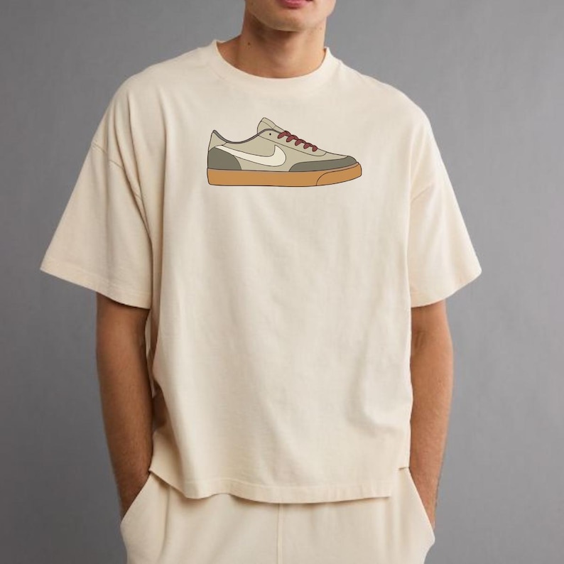 May include: A cream-colored short-sleeve t-shirt featuring a centered graphic illustration of a low-top sneaker. The sneaker design displays a light beige upper, a white swoosh logo, dark red laces, and a solid gum-colored rubber sole.