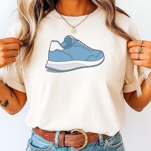 May include: A cream-colored short-sleeve t-shirt featuring a graphic illustration of a light blue sneaker with a white heel tab and a thick white sole. The sneaker is positioned centrally on the chest area of the shirt, showcasing a side profile.