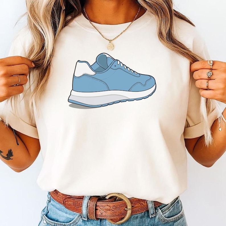 May include: A cream-colored short-sleeve t-shirt featuring a graphic illustration of a light blue sneaker with a white heel tab and a thick white sole. The sneaker is positioned centrally on the chest area of the shirt, showcasing a side profile.