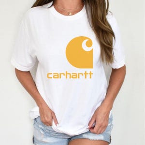 May include: A white short-sleeved t-shirt featuring a large yellow graphic of the Carhartt logo on the chest. The logo consists of a stylized C shape above the word carhartt printed in a bold, lowercase, sans-serif yellow font.