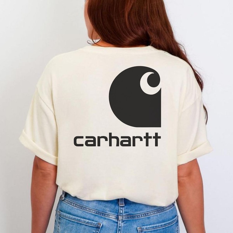 May include: A cream-colored short-sleeve t-shirt featuring a large black Carhartt logo printed on the back. The graphic includes the iconic stylized C emblem positioned above the brand name text in a bold, sans-serif black font.