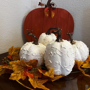 May include: Three white pumpkins with raised, swirling white texture patterns and dark brown stems, arranged on a glass surface with artificial orange and yellow autumn leaves and small berry clusters, with a large flat wooden pumpkin behind.