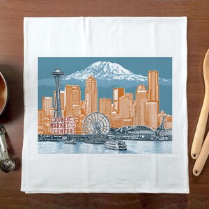 May include: White kitchen towel featuring a colourful illustration of the Seattle skyline, including the Space Needle, and the text "Public Market Center".