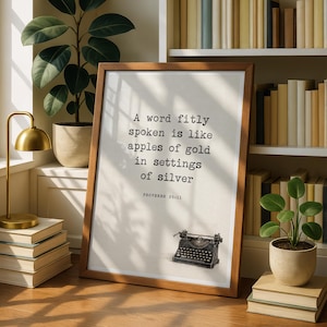 May include: A framed wall art print featuring the text A word fitly spoken is like apples of gold in settings of silver, followed by PROVERBS 25:11. Below the text is a small illustration of a black vintage typewriter on a light background.