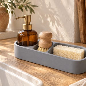 Op de afbeelding: Een grijs ovaal gootsteenorganizer-bakje met een bruine glazen zeepdispenser met gouden pomp, een ronde houten afwasborstel met lichte haren en een rechthoekige beige cellulosespons voor het reinigen en organiseren van de keuken.