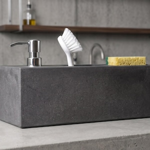 May include: A rectangular dark grey concrete sink caddy featuring a built-in silver metal soap dispenser pump, a white plastic dish brush with stiff bristles, and a yellow rectangular kitchen sponge resting in a dedicated storage compartment.