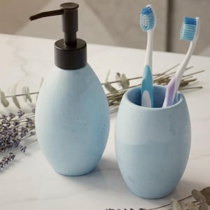 May include: A light blue ceramic bathroom set featuring a soap dispenser with a black pump and a toothbrush holder with two toothbrushes. The set has a speckled finish and a modern design. The dispenser is egg-shaped.
