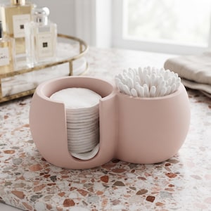 May include: A matte light pink dual-compartment bathroom organiser. One side features a vertical cutout holding a stack of round white cotton pads, while the other side holds a cluster of white cotton swabs with paper stems in an upright position.