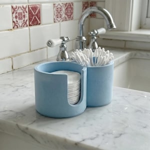 Può includere: Un organizer da bagno in cemento blu chiaro con due scomparti. Uno contiene dischetti di cotone, l'altro cotton fioc. L'organizer è posizionato su un piano di lavoro in marmo vicino a un lavandino e un rubinetto.