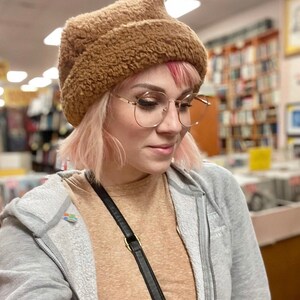 Puede incluir: Una mujer con el pelo rosa lleva un gorro de pelo marr&oacute;n y gafas de montura dorada. Lleva una sudadera gris sobre una camisa marr&oacute;n.