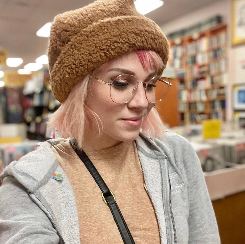 Puede incluir: Una mujer con el pelo rosa lleva un gorro de pelo marr&oacute;n y gafas de montura dorada. Lleva una sudadera gris sobre una camisa marr&oacute;n.