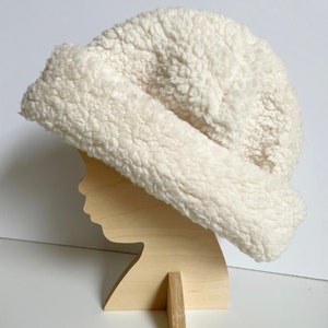 May include: A white fluffy hat made of faux fur. The hat is on a wooden mannequin head.