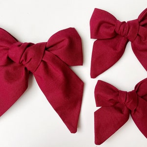 May include: Three burgundy fabric hair bows of different sizes. The bows are tied with a knot in the center and have pointed ends.