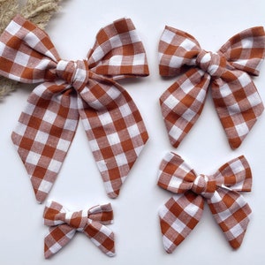 May include: Set of four brown and white gingham hair bows in different sizes. The bows are made of fabric and have a checkered pattern.