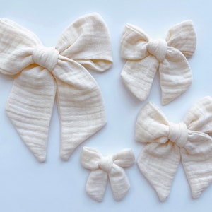 May include: Four off-white fabric hair bows of varying sizes. The bows are made of a soft, textured fabric and are tied in a simple knot.