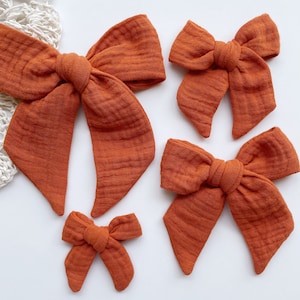 May include: Four burnt orange fabric hair bows of varying sizes. The bows are made of a soft, textured fabric and are tied with a simple knot in the center.