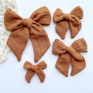 May include: Four rust-colored fabric bows of varying sizes are arranged on a white surface. The bows have a textured appearance and are tied in the center. The largest bow has long, flowing ends, while the smallest is a simple knot.